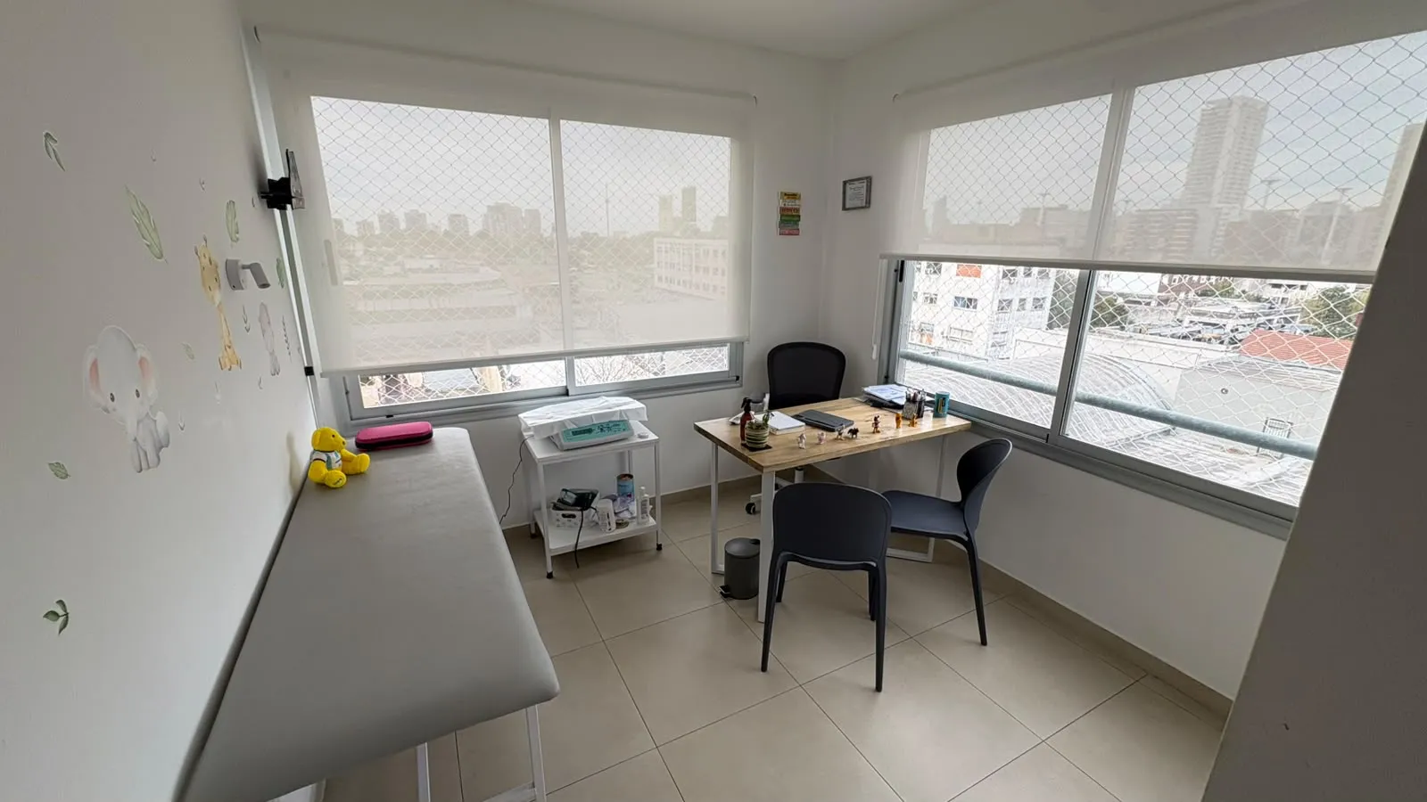 Consultorio con una camilla blanca con un osito arriba, una mesa de trabajo de madera junto a dos ventanales y calcomanías de una jirafa y un elefante en la pared.