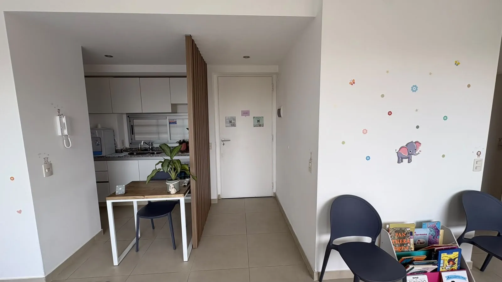 Recibidor del consultorio, con la puerta de entrada, una cocina pequeña detrás de un panel de listones de madera y una pared blanca con calcomanías de flores y un elefante.