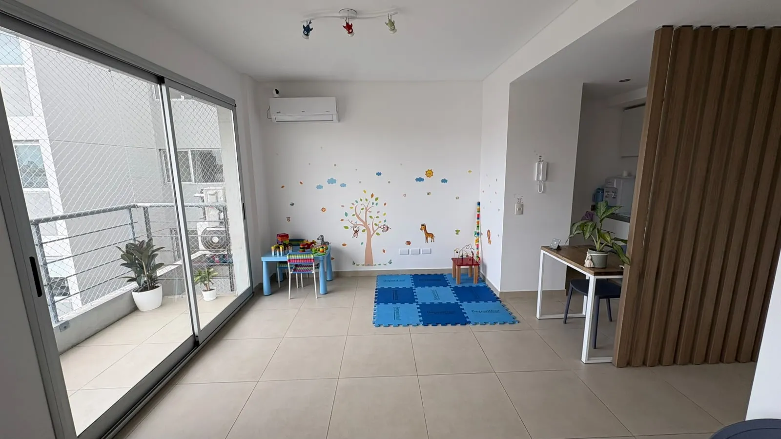 Rincón de juegos con una alfombra de goma azul en el piso, una mesita celeste con sillas y juguetes, y un árbol dibujado en la pared con monitos y una jirafa.