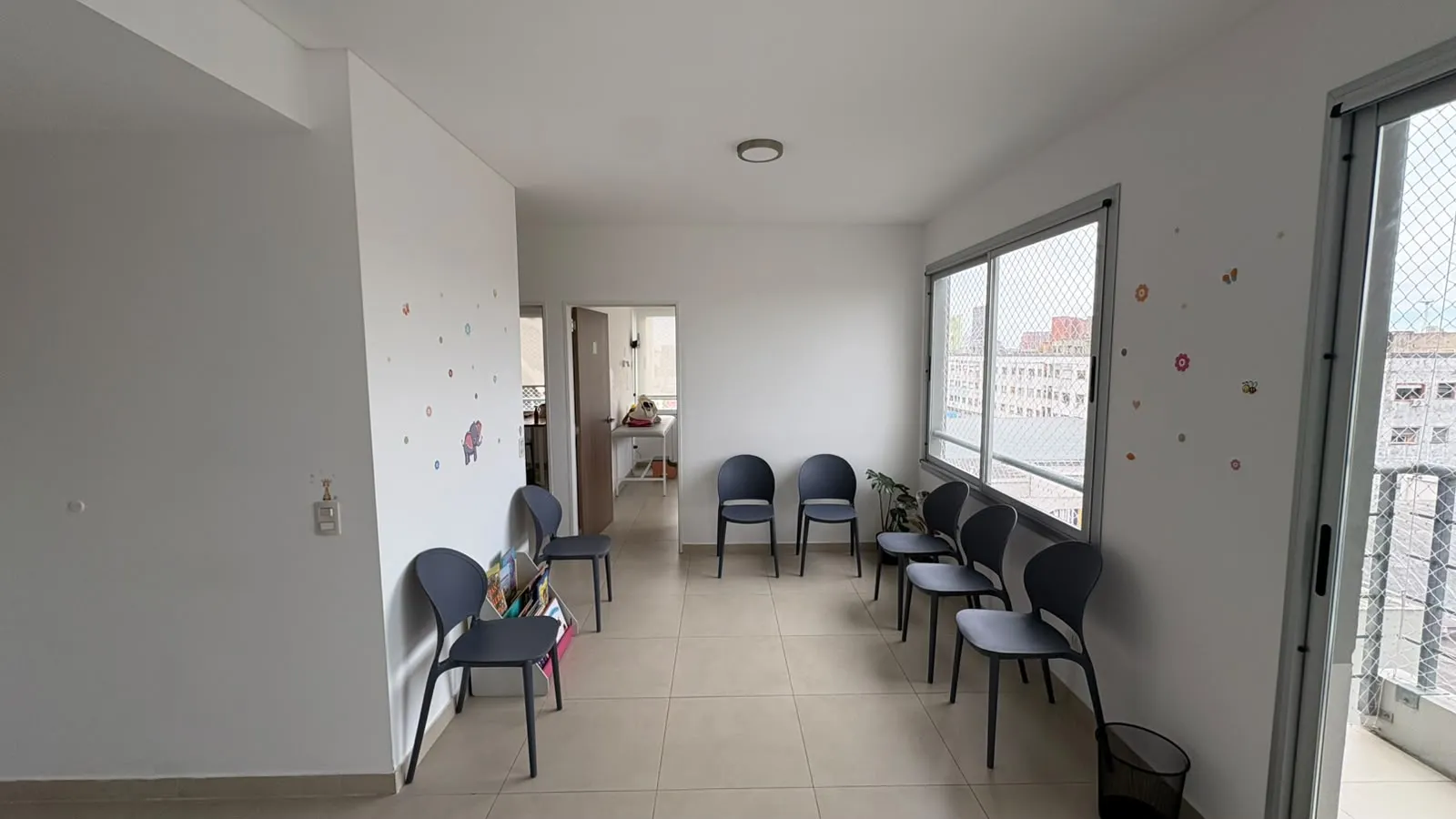 Sala de espera con siete sillas oscuras contra las paredes, un ventanal con red de protección, un revistero con libros infantiles y calcomanías de mariposas y flores en la pared.