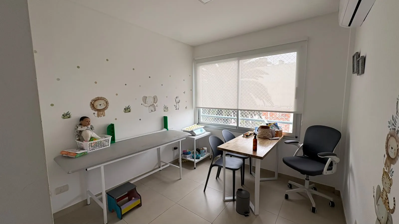 Segundo consultorio con una camilla larga, una balanza para bebés sobre una mesa baja, un escritorio con sillas y calcomanías de un león, un elefante y una cebra en la pared.